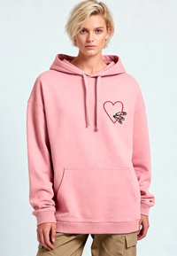 Felpa rosa realizzata in tessuto morbido, dotata di tasca frontale e cuore ricamato con design di spilla di sicurezza nell'area sinistra del petto.
