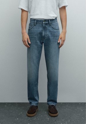 Personne portant un jean bleu droit, un t-shirt blanc et des chaussures à lacets marron foncé debout sur un sol gris.