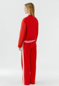 Frau steht, Blick nach links, trägt eine rote Jacke und passende weit geschnittene Hosen mit weißen Seitenstreifen vor einem schlichten weißen Hintergrund.