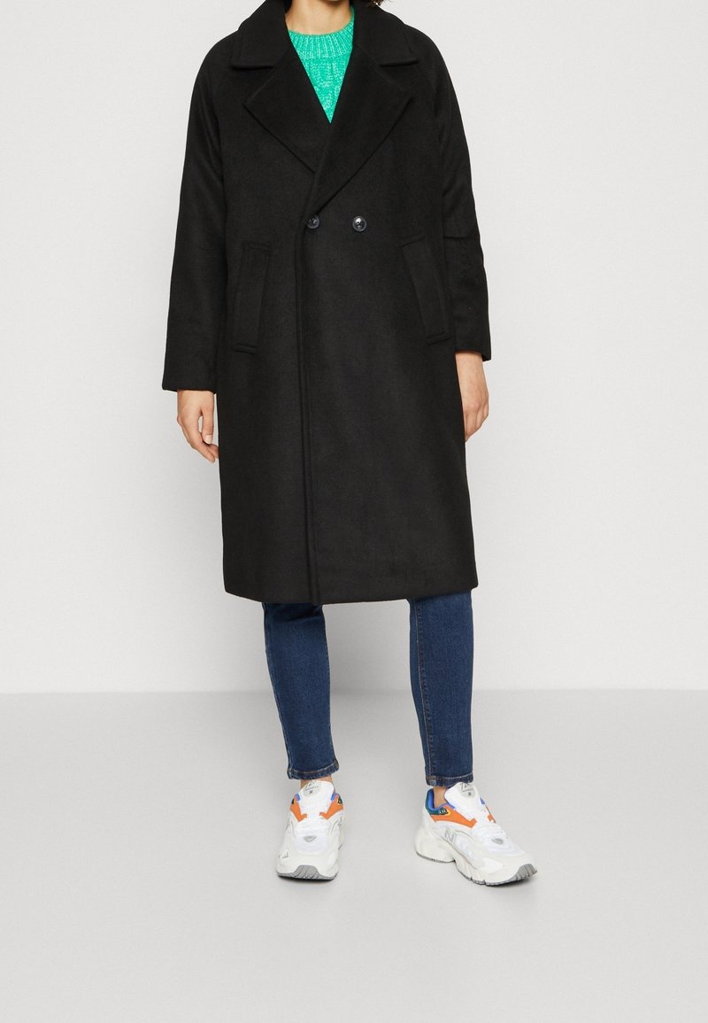 Personne portant un manteau long noir à double boutonnage, un jean bleu, des baskets blanches avec des détails orange et bleu, et un pull en maille vert.