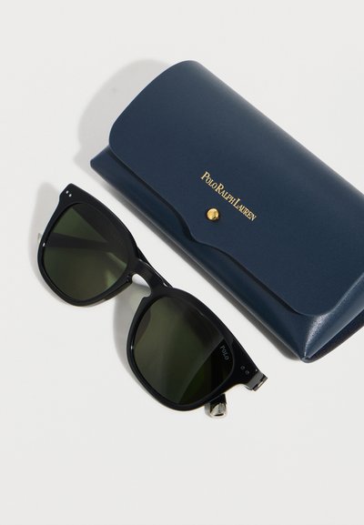 Μαύρα ορθογώνια γυαλιά ηλίου με σκοτεινούς φακούς δίπλα σε θήκη Polo Ralph Lauren σε ναυτικό μπλε με κλείσιμο σε χρυσό κουμπί.