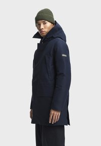 Marineblauwe parka met een hoge kraag, capuchon en zijzakken. Beschikt over een logopatch op de linkerarm en een strakke, gladde stofstructuur.