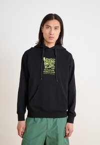 Black sweatshirt with hood, featuring a yellow graphic design and text. Made of cotton, it has a front pocket and ribbed cuffs. Model wears green shorts.