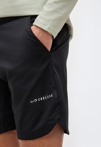 Personne portant un short noir avec le texte "ENDLESS" et un symbole d'infini sur la jambe, main dans la poche, et une chemise à manches longues à texture légère.
