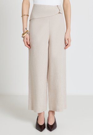 Pantaloni beige a gamba larga con cintura asimmetrica e chiusure frontali con ganci, abbinati a tacchi scuri con punta affilata e braccialetti dorati al polso sinistro.