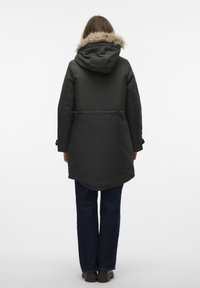 Parka nera con cappuccio foderato in pelliccia, caratterizzata da una silhouette leggermente sfiancata, maniche lunghe e regolatori laterali, abbinata a jeans blu e scarpe scure.