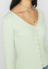 Ljusgrön ribbad cardigan med V-hals, pärlknappar framtill och en strukturerad tyg. Lager av guldkedjor.