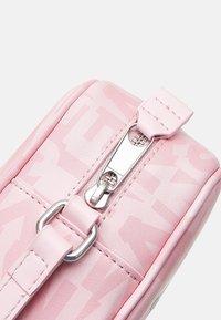 Różowa skórzana torba o błyszczącej fakturze, z wyraźnym kontrastowym nadrukiem logo, srebrnym zamkiem błyskawicznym oraz pasującym różowym paskiem z detalami w postaci przeszyć.
