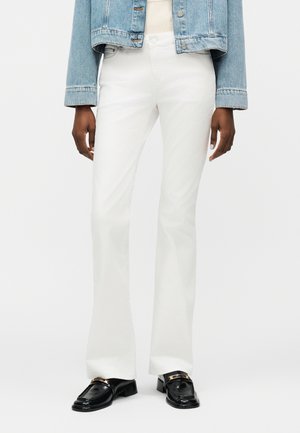 Isik, kes kannab valgeid sirge säärega pükse, musti kuldsete pandlatega loafer-sussisid, beeži topi ja lühemaks jäänud helesinist teksajopet.