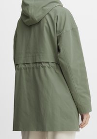 Giacca verde oliva con cappuccio, dal taglio morbido, caratterizzata da una vita arricciata e maniche lunghe. Il materiale appare testurizzato e leggero.
