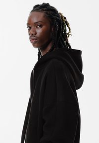 Felpa nera con zip e fit rilassato, dotata di un cappuccio ampio e una texture liscia, indossata da una persona con lunghi capelli intrecciati.