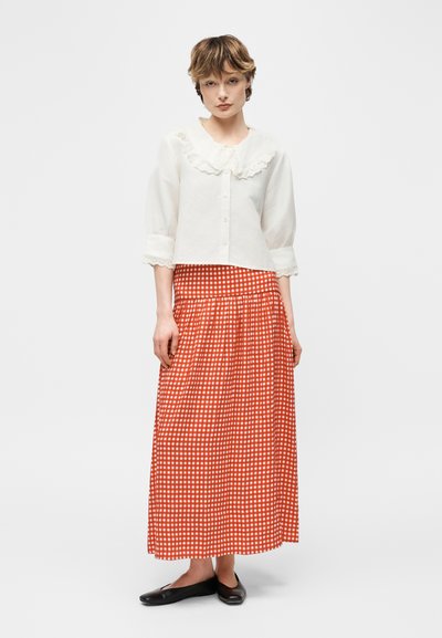 Νεαρή γυναίκα που φοράει λευκό πουκάμισο με βολάν στη γιακά, πορτοκαλί και λευκή καρό μακριά φούστα και μαύρα μπαλέτα, στέκεται σε απλό φόντο.