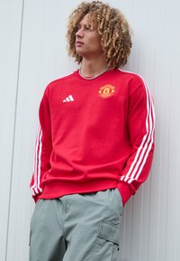 Rode sweatshirt met witte strepen op de mouwen en het Manchester United-logo, gecombineerd met lichtgrijze cargobroek. Eenvoudige ronde halslijn.
