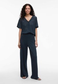 Jūras zils kombinezons ar īsām piedurknēm, V veida kakla izgriezumu un platām biksēm. Ar zigzaga rakstu un brīvu piegriezumu, izgatavots no viegla materiāla.