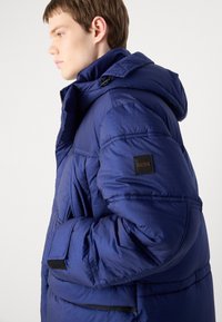 Giubbotto imbottito navy con cappuccio, caratterizzato da una texture trapuntata, tasche laterali e una patch in pelle nera con logo sulla manica.