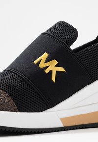 Baskets noires à enfilage avec maille respirante, semelle blanche, capuchon marron et logo doré MK sur la bande élastique en haut.
