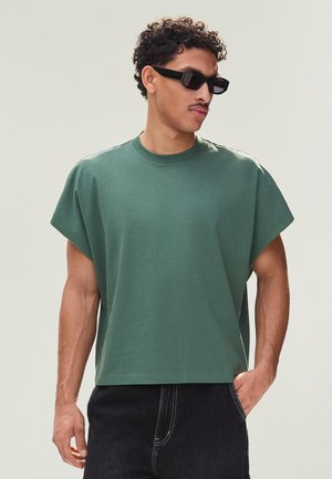 Man wearing dark sunglasses, green short-sleeve oversized T-shirt, and black pants, standing with one hand in pocket, looking sideways.