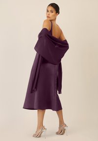 Lila Satinrobe mit Spaghettiträgern, einem tiefen Rücken und einem fließenden Saum, kombiniert mit einem transparenten Schal und silbernen High-Heels.
