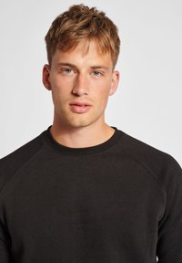 Sort sweatshirt med rund halsudskæring, raglanærmer og en blød tekstur. Modellen har kort hår og en neutral ansigtsudtryk.