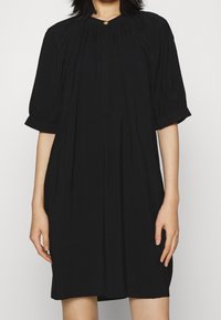 Svart klänning med korta puffärmar, rund halsringning, vertikala veck och en liten metallknapp som detalj vid halsen. Slät tygtextur.