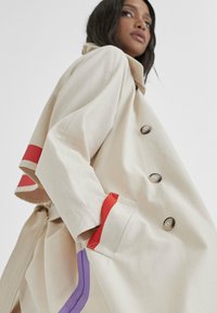 Beżowy płaszcz trench z dużymi guzikami, czerwonymi i fioletowymi akcentami na rękawach oraz paskiem. Wykonany z teksturowanej tkaniny, charakteryzujący się luźnym krojem i bocznymi kieszeniami.