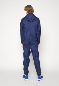 Ναυτικό μπλε αδιάβροχο με κουκούλα, ελαστικούς σφιγκτήρες, και ταιριαστά παντελόνια, με λεία, ανακλαστική υφή και χαλαρή εφαρμογή.