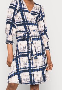 Femme portant une robe portefeuille mi-longue à motifs à carreaux bleu, noir et blanc, avec des manches trois-quarts bouffantes et des poignets à volants.