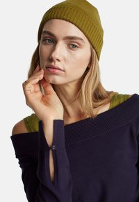 Námořnický pletený svetr s designem s odkrytými rameny a žebrovaným výstřihem. Olivově zelené tričko je viditelné u výstřihu. Čepice je olivově zelená.