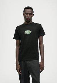 Μαύρο βαμβακερό T-shirt με κοντά μανίκια, με πράσινο графικό λογότυπο στην μπροστινή πλευρά. Φορεμένο με σκούρες γκρι παντελόνες, το μοντέλο στέκεται κατά πρόσωπο.