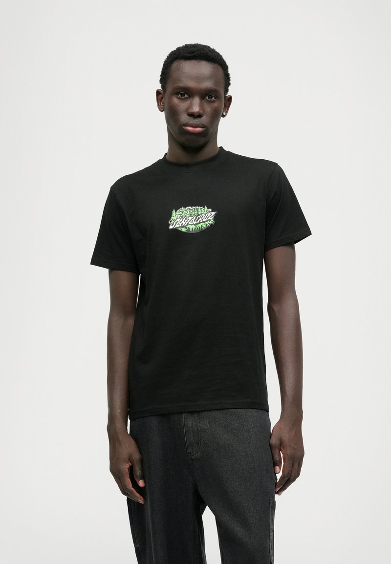 Μαύρο βαμβακερό T-shirt με κοντά μανίκια, με πράσινο графικό λογότυπο στην μπροστινή πλευρά. Φορεμένο με σκούρες γκρι παντελόνες, το μοντέλο στέκεται κατά πρόσωπο.