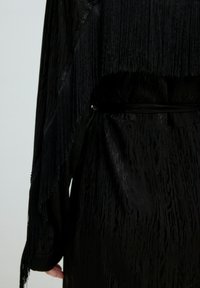 Schwarzes Kleidungsstück aus strukturiertem Stoff mit einem Fransen-Detail auf einer Seite, tailliert mit einem Gürtel, mit langen Ärmeln und einem mehrlagigen Design.