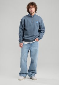 Gray half-zip sweatshirt with blue flower design, worn over a white t-shirt, paired with light blue wide-leg jeans and white sneakers.
