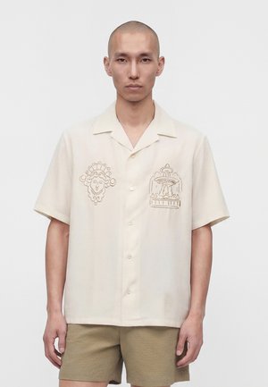 Homme à la tête rasée portant une chemise beige à manches courtes boutonnée avec des motifs brodés de visages et de fontaines, assortie à un short marron clair.
