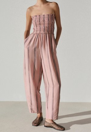 Frau trägt einen trägerlosen pinken Jumpsuit mit vertikalen schwarzen Streifen und Händen in den Taschen, kombiniert mit schwarzen Netz-Ballerinas.