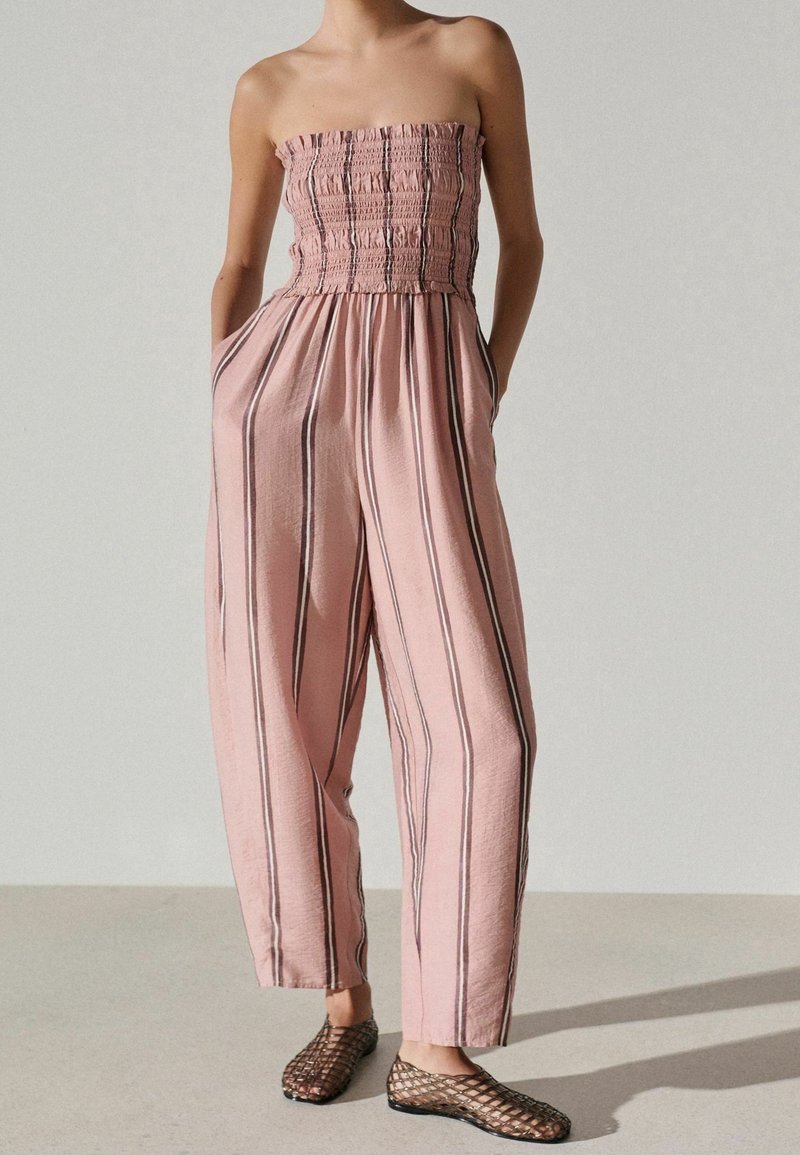 Femme portant une combinaison bustier rose à rayures noires verticales avec les mains dans les poches, assortie à des chaussures plates noires en résille.