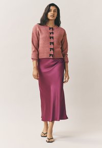 Roza pletena cardigan s črnimi zaponkami v obliki pentlje, kombinirana z dolgo, gladko, satensko vijolično krilo. Črne japonke z naramnicami dopolnjujejo videz.