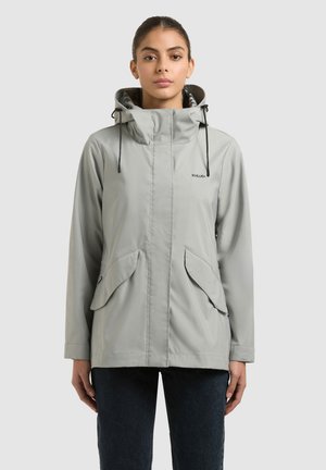 Žena v svetlosivej bundi s kapucňou, prednými vreckami so záklopkou a zapínaním na zips, stojaca pred jednoduchým svetlým pozadím.