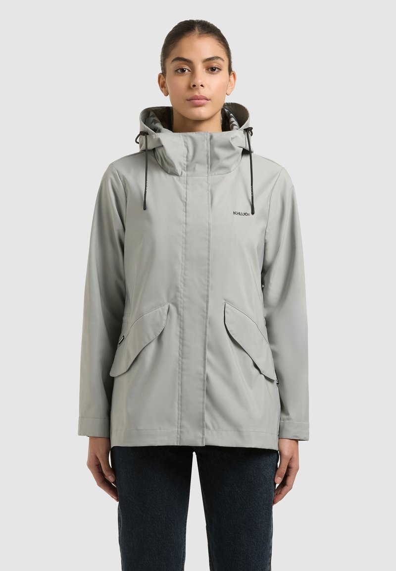 Žena v svetlosivej bundi s kapucňou, prednými vreckami so záklopkou a zapínaním na zips, stojaca pred jednoduchým svetlým pozadím.