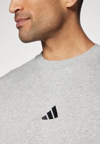 Sweatshirt de algodão cinza com decote redondo e um logótipo preto da Adidas com três riscas no peito. Uma textura subtil é visível.