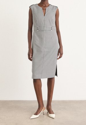 Sukienka bez rękawów do kolan w kratę w jodełkę z paskiem, rozcięciem z przodu i białymi, szpiczastymi sandałkami typu slingback na stojącej osobie.