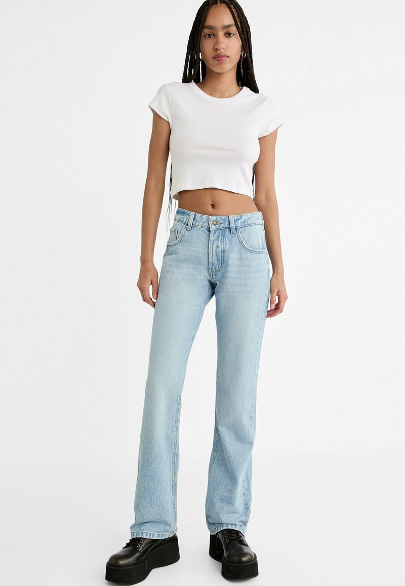 Jeans azzurri chiari a vita alta con design a gamba dritta, indossati con una t-shirt bianca corta. Scarpe con zeppa nere completano il look.