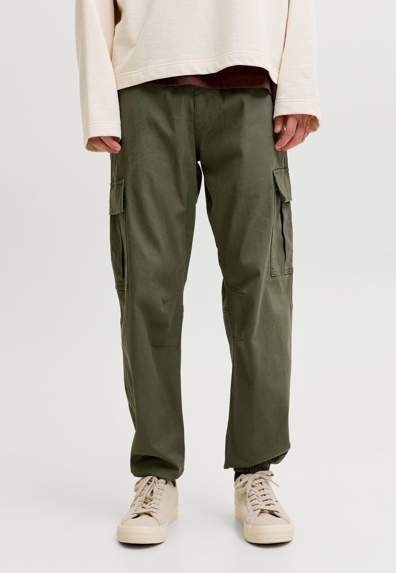 Personne portant un pantalon cargo vert olive, des baskets beiges et un haut à manches longues crème, debout sur un fond clair uni.