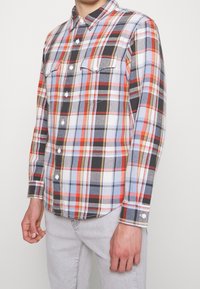 Geruit flanellen overhemd met een mix van rode, blauwe en zwarte kleuren, knoopsluiting aan de voorkant, kraag en twee borstzakken met kleppen.