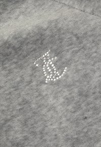 Sivi materijal s mekom teksturom s dekorativnim logotipom izrađenim od sjajnih rhinestona u stiliziranom dizajnu.