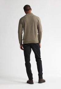 Pulover verde măsliniu cu guler înalt, design ajustat; asortat cu blugi negri și pantofi maro. Textura pare netedă și tricotată.