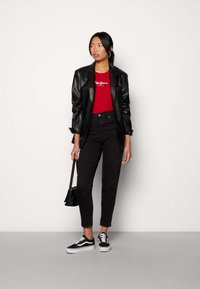 Blazer in finta pelle nera sopra una maglietta rossa aderente con logo bianco, jeans neri e sneakers nere con dettagli bianchi. Piccola borsa nera.