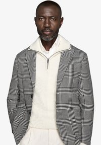 Blazer a scacchi bianco e nero con un pullover bianco testurizzato, dotato di colletto con zip, indossato sopra una camicia bianca. Vestibilità su misura.