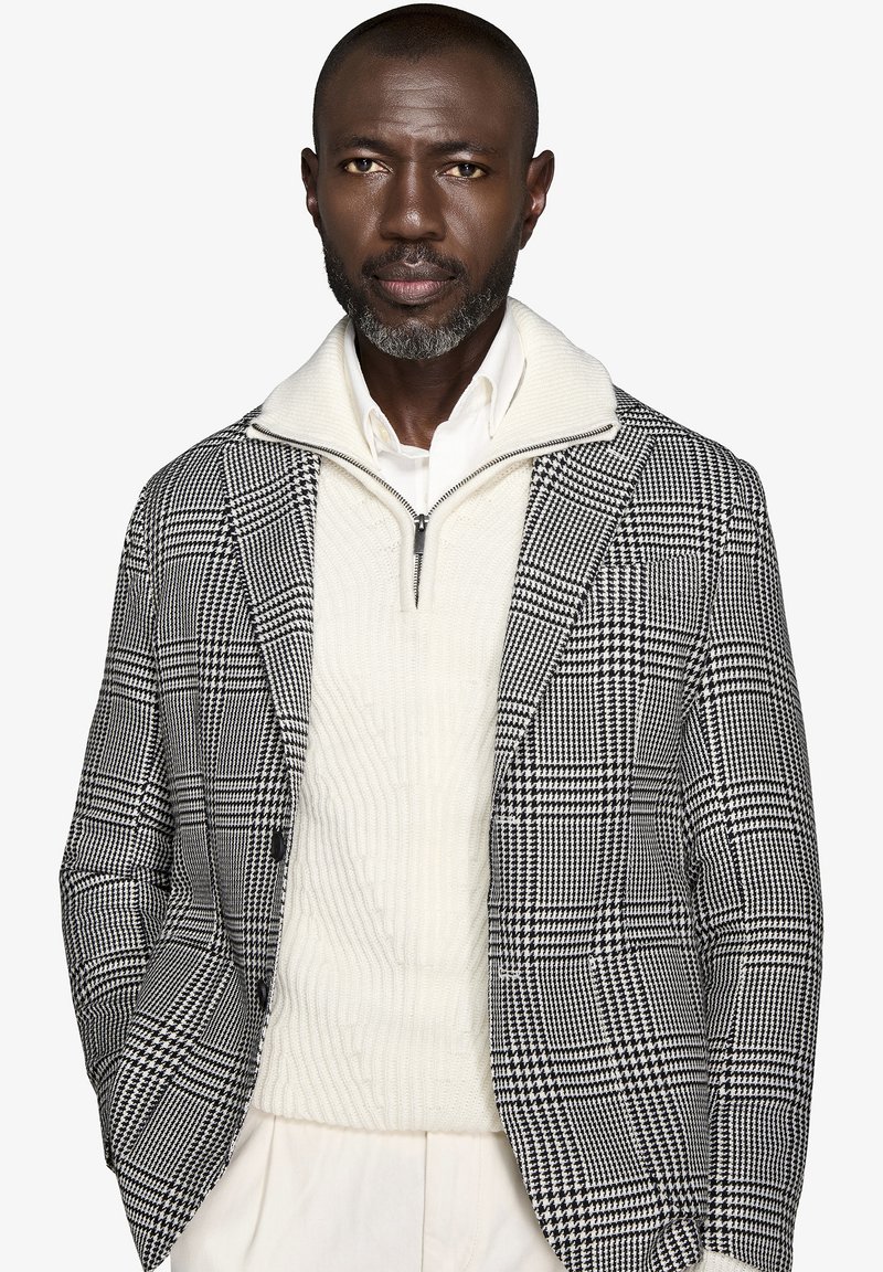 Blazer a scacchi bianco e nero con un pullover bianco testurizzato, dotato di colletto con zip, indossato sopra una camicia bianca. Vestibilità su misura.
