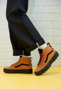 Bruna mocka hög-top sneakers med svarta accenter och röda snören. Har vadderad krage, texturerad sula och kontrasterande sömmar.