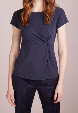 Vrouw in een marineblauw topje met korte mouwen en een knoop aan de zijkant, gecombineerd met donkerblauwe geruite broek, tegen een effen achtergrond.